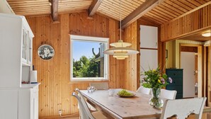 Interior de madera con una mesa de comedor, sillas, un jarrón con flores y una ventana que da a vistas de vegetación.