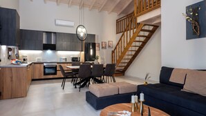 A modern kitchen with dark cabinets, a dining area with a wooden table and chairs, a staircase, and a living room with a sofa and a coffee table.