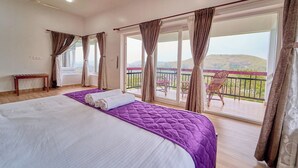 A bedroom with a large bed, a balcony with chairs, and a view of the landscape. - Priyas Villa Vagamon by VOYE HOMES (Peermade)