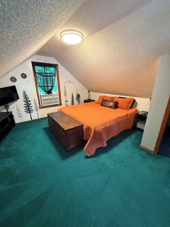 A bedroom with a sloped ceiling, a bed with an orange bedspread, a wooden bench, a TV, and a window with a view of trees.