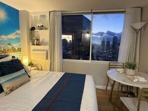 Une chambre d’hôtel comprenant un lit, une table à manger, une chaise, une fenêtre donnant sur la ville et un mur orné d’une fresque aux motifs tropicaux.
