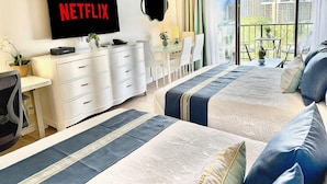 Habitación de hotel con un televisor grande mostrando 'NETFLIX', un escritorio con una silla, una cama con ropa de cama azul y blanca, y un balcón con vistas a la ciudad.