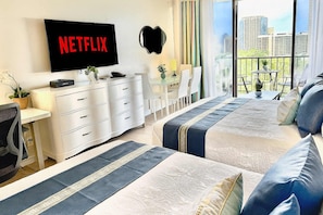 Quarto de hotel com uma televisão grande exibindo 'NETFLIX', uma escrivaninha com cadeira, uma cama com roupa de cama azul e branca, e uma varanda com vista para a cidade.