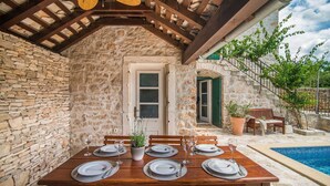 A stone-clad dining area with a wooden table set for six, a stone wall, and a view of a swimming pool. - Villa Ivana Summary: (Mirca)