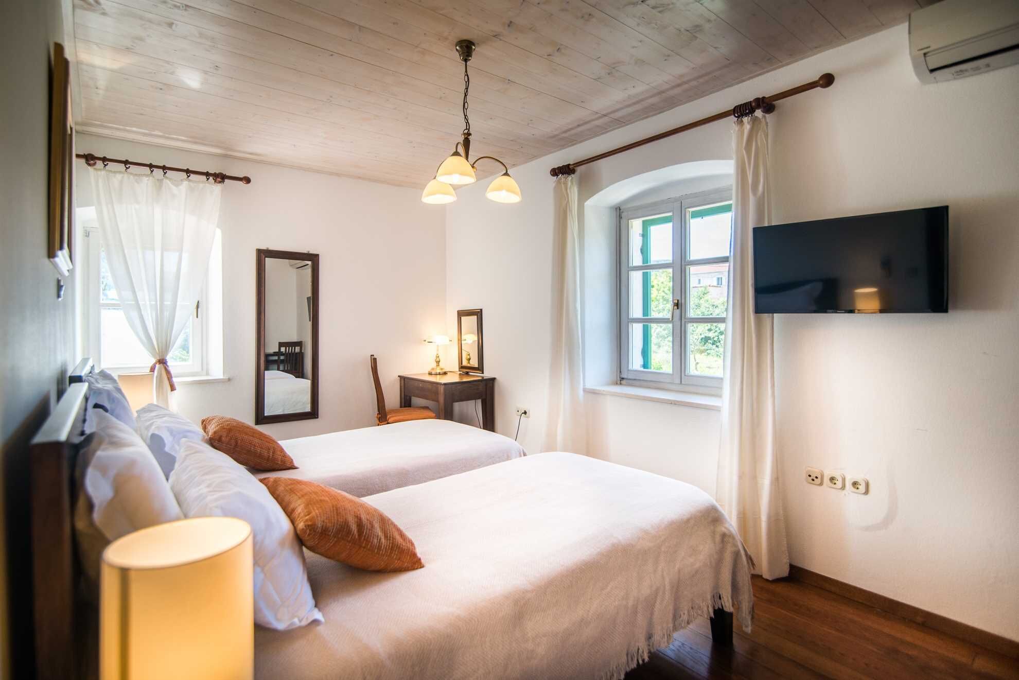 A bedroom with two beds, a mirror, a desk with a lamp, a TV mounted on the wall, and a window with curtains.