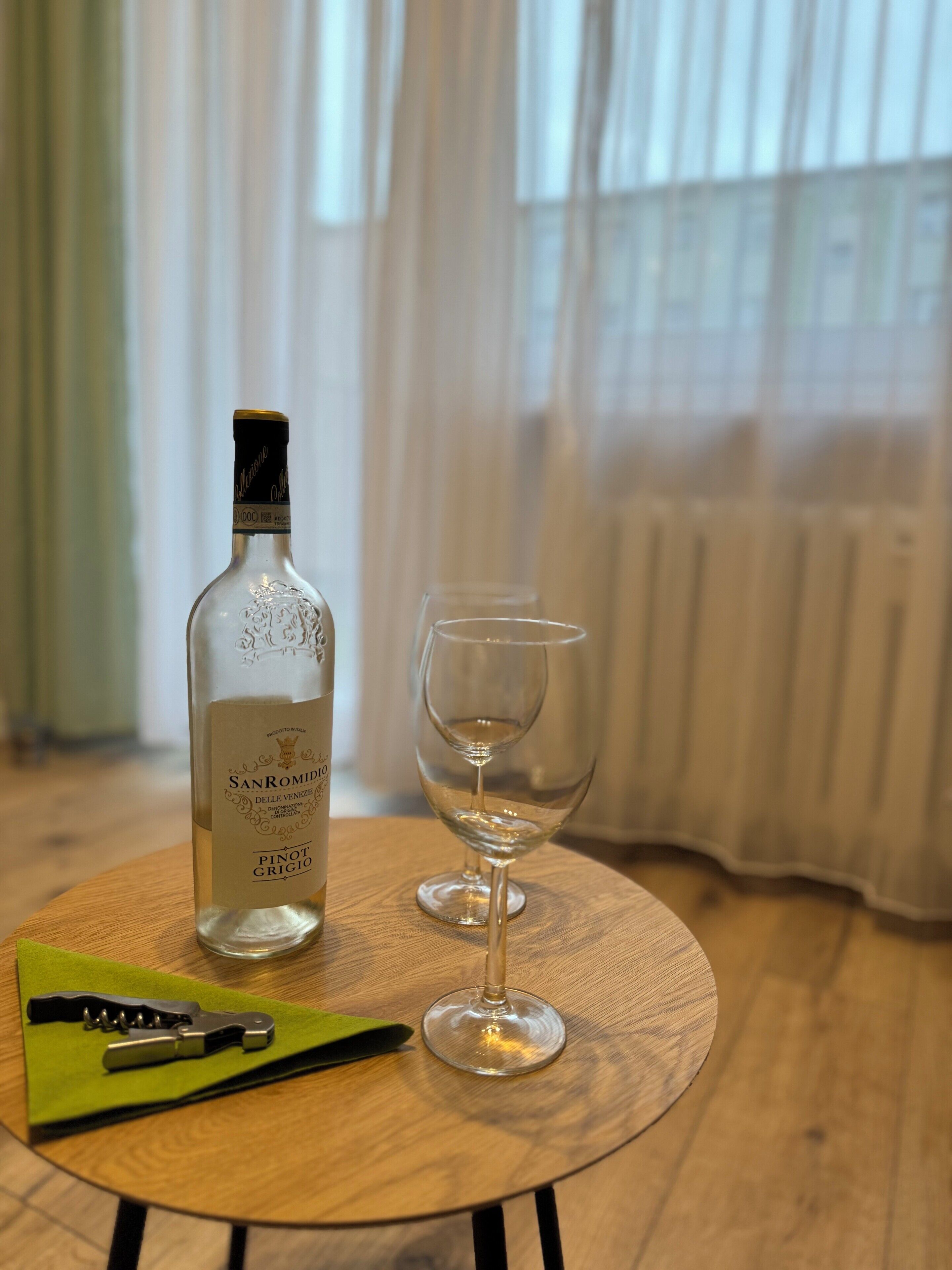 A bottle of San Romido Pinot Grigio wine and an empty wine glass on a round wooden table in a room with a window and curtains.