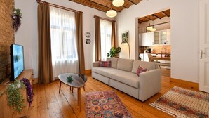 A cozy living room with a sofa, a coffee table, and a TV. There's a kitchen in the background with a dining area.