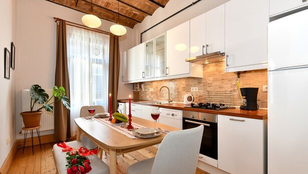 A modern kitchen with white cabinets, a wooden countertop, and a built-in oven. There is a dining table with two chairs, a potted plant, and a vase with red roses.