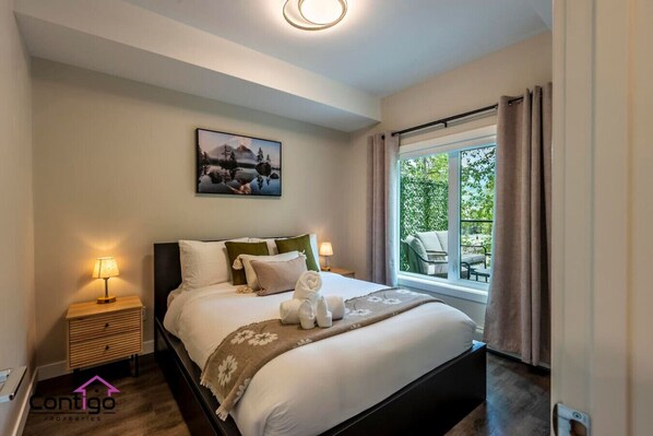 A bedroom with a bed, a nightstand, a lamp, a window with curtains, and a painting on the wall.