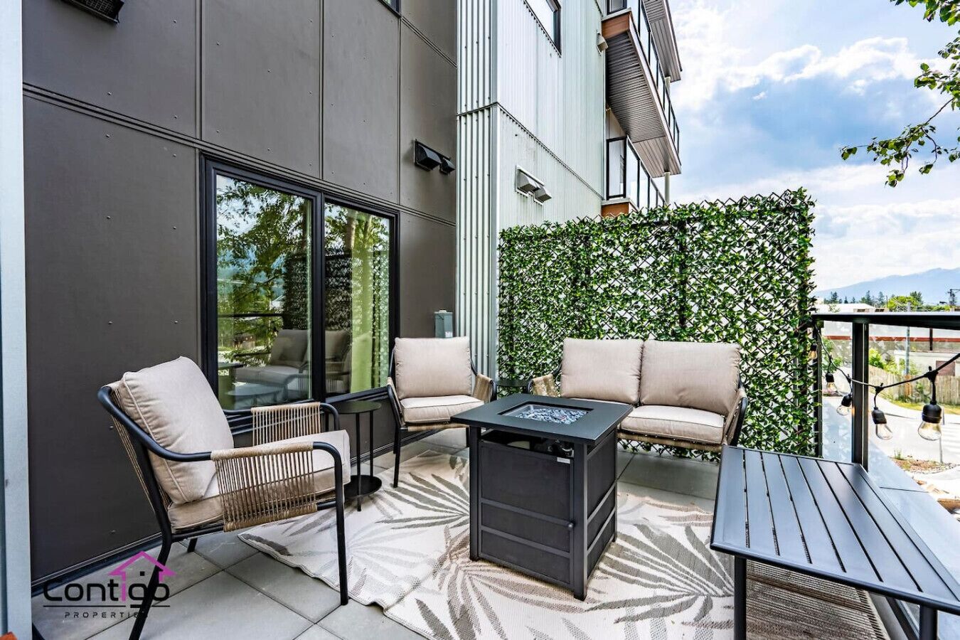Une terrasse extérieure avec un canapé, des fauteuils et une table, entourée d’une haie et d’un mur de verre.