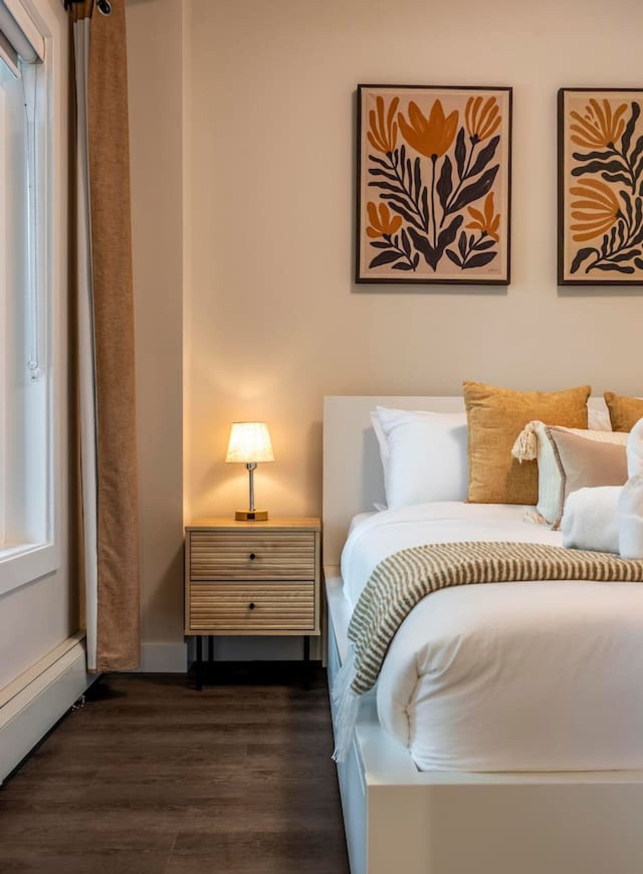 Une chambre à coucher avec un lit, une table de chevet, une lampe et une œuvre d’art accrochée au mur.