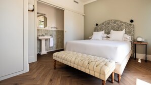 Een slaapkamer met een groot bed, een bankje, een nachtkastje en een badkamer met een wastafel en spiegel.
