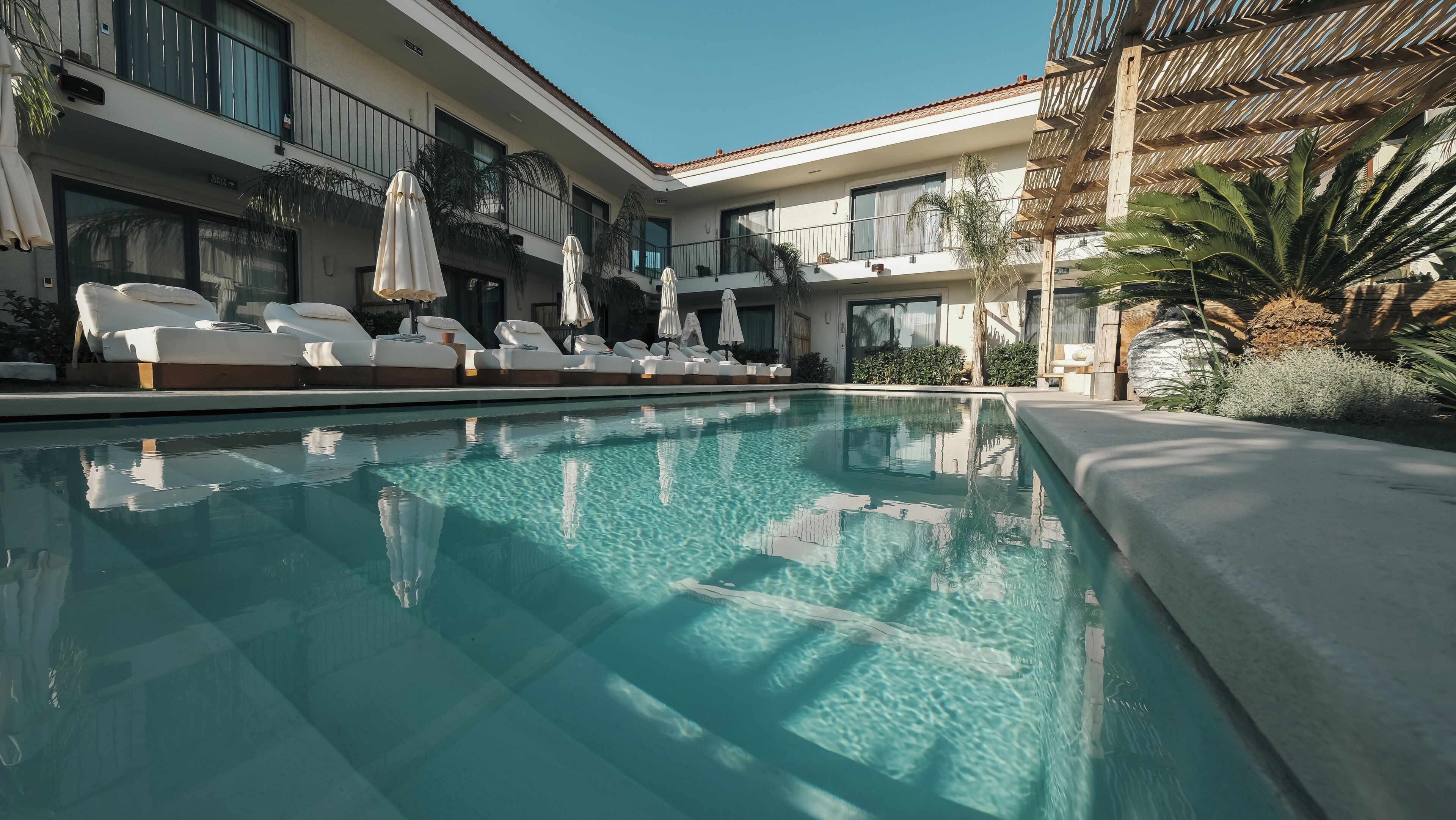 A swimming pool with lounge chairs and umbrellas, surrounded by a building with balconies and plants.