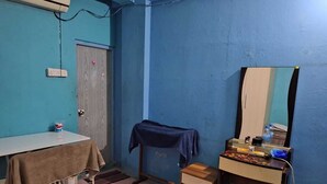 A small room with a blue wall, a white table with a brown cloth, a wooden stool, a small wooden nightstand, and a mirror on a wooden dresser.
