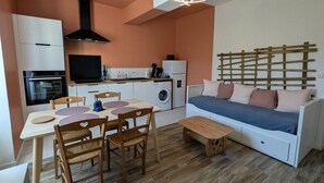 Een knus woongedeelte met een kitchenette, een eethoek met tafel en stoelen, een bank met kussens en een wasmachine.