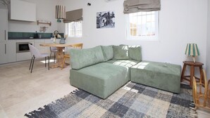 A modern apartment with a green sectional sofa, a dining area, a kitchen with white cabinets, and a large window with a view.