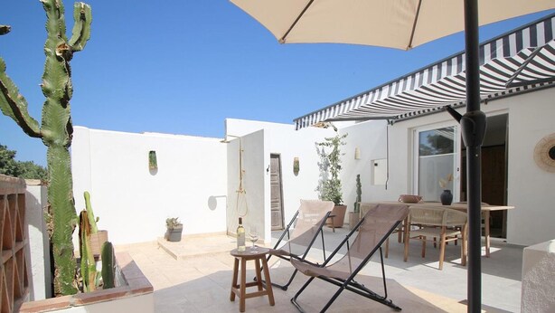A patio with a chair, table, and umbrella, surrounded by cacti and a white building.