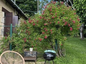 Un jardin agrĂ©mentĂ© dâun arbuste aux fleurs rouges, dâune chaise en osier, dâun barbecue et dâun bĂątiment au style rustique.