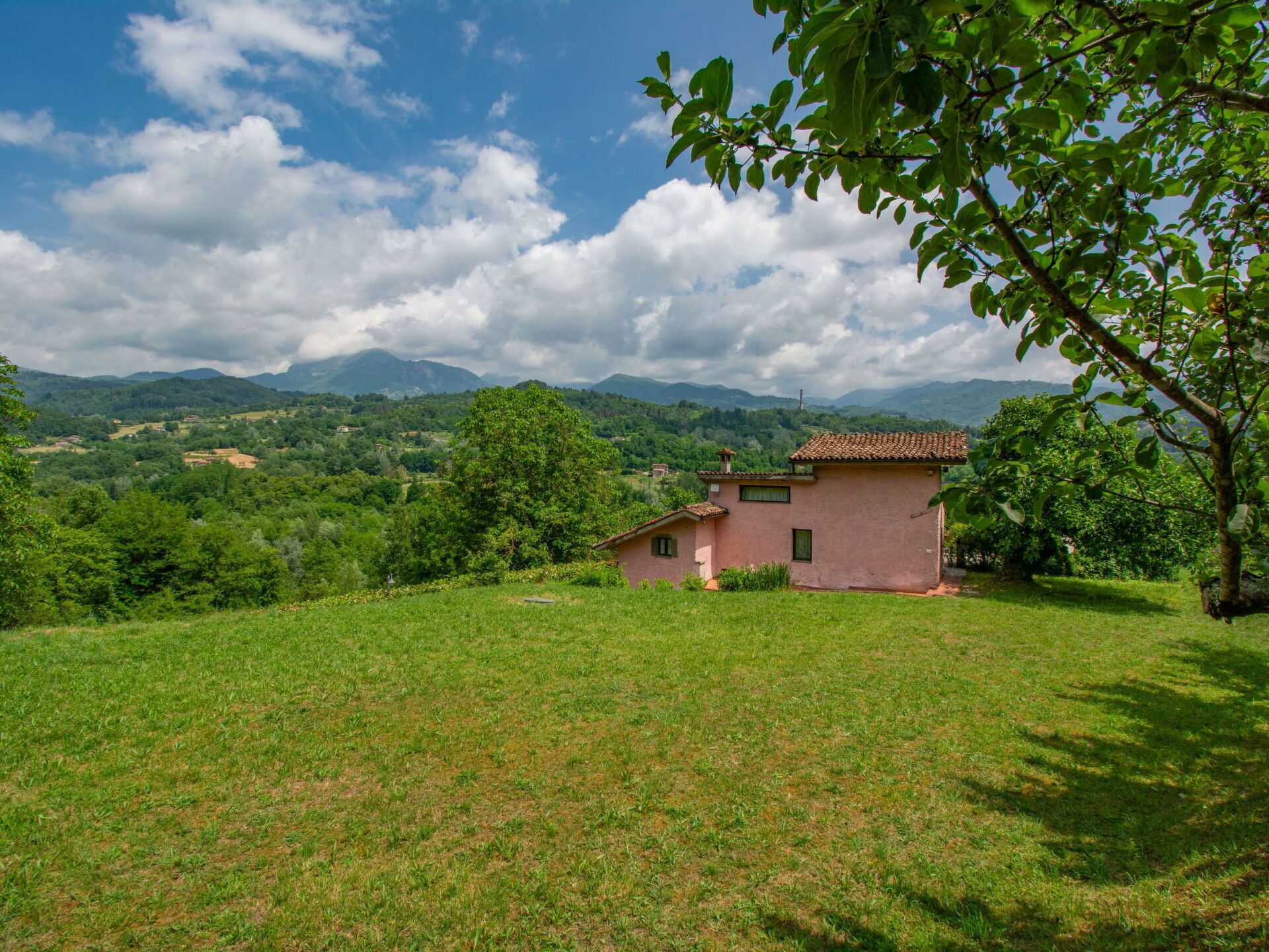 Une maison de plain-pied, avec un toit en tuiles, entourée d’une pelouse verdoyante et d’arbres, se dressant sur un fond de collines douces et sous un ciel partiellement nuageux.