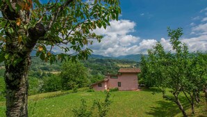 Una casa rustica con tetto in tegole, circondata da campi verdi rigogliosi e alberi, con montagne e un cielo parzialmente nuvoloso sullo sfondo.
