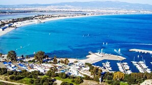 Eine Vogelperspektive auf eine Küstenmarina mit vertäuten Booten, einem Sandstrand und einer Stadtlandschaft im Hintergrund.