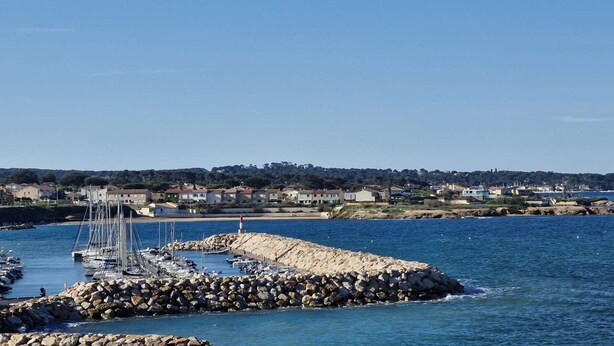 Un port de plaisance situé sur la côte, avec des bateaux amarrés, une digue rocheuse et une zone résidentielle en arrière-plan.