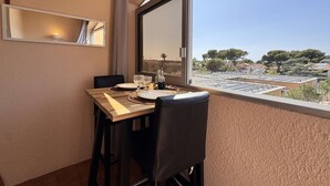 Un coin repas avec une table dressée pour deux personnes, une fenêtre donnant sur des bâtiments et des arbres, et un miroir fixé au mur.
