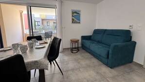 Una sala de estar moderna con un sofá azul verdoso, una mesa de comedor con sillas y un balcón con vistas a edificios y zonas verdes.