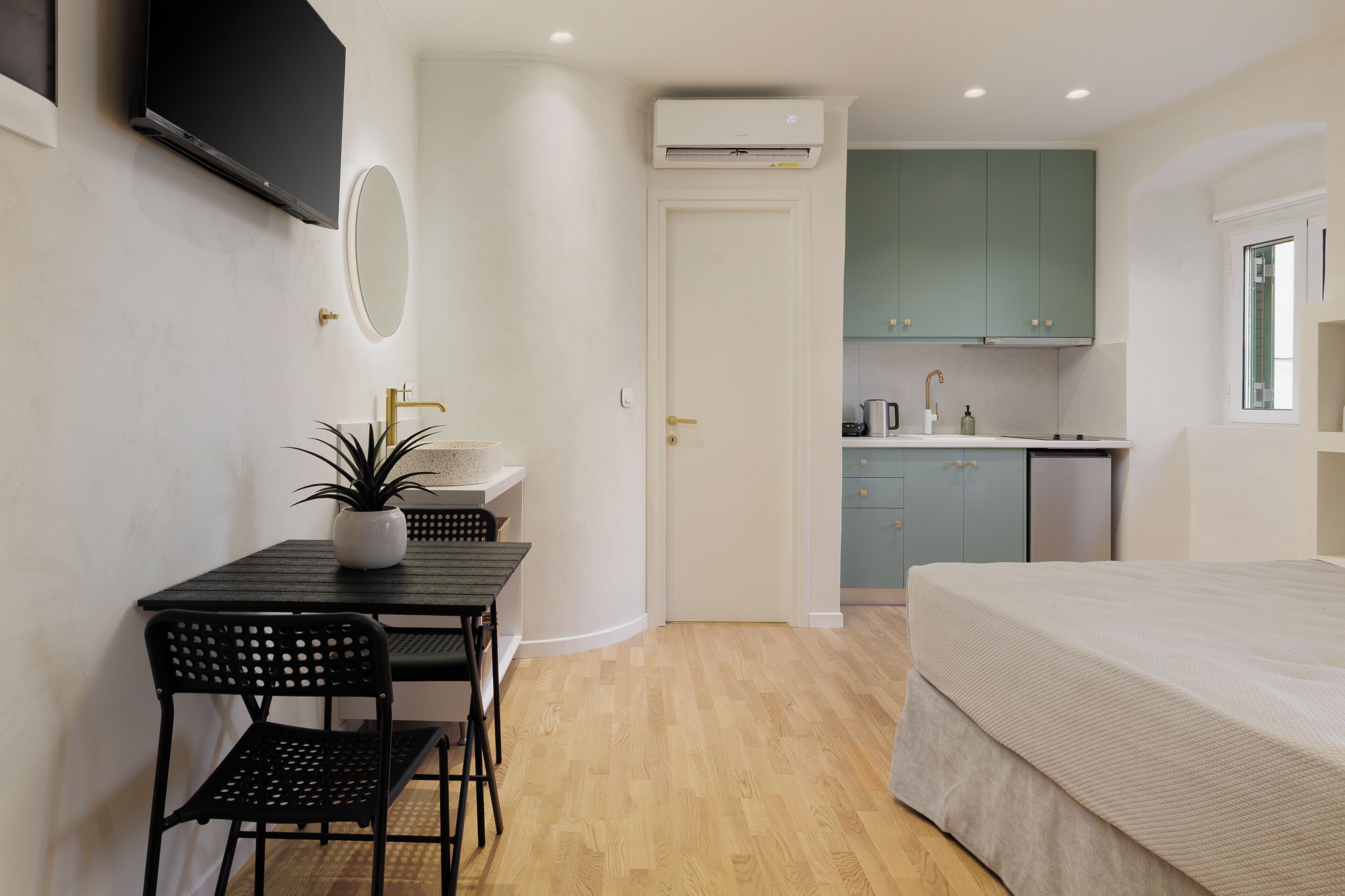 Een compacte kamer met een bed, een eettafel met stoelen, een potplant, een aan de muur bevestigde televisie, een keukenhoek met kastjes en een spoelbak, en een airconditioning.