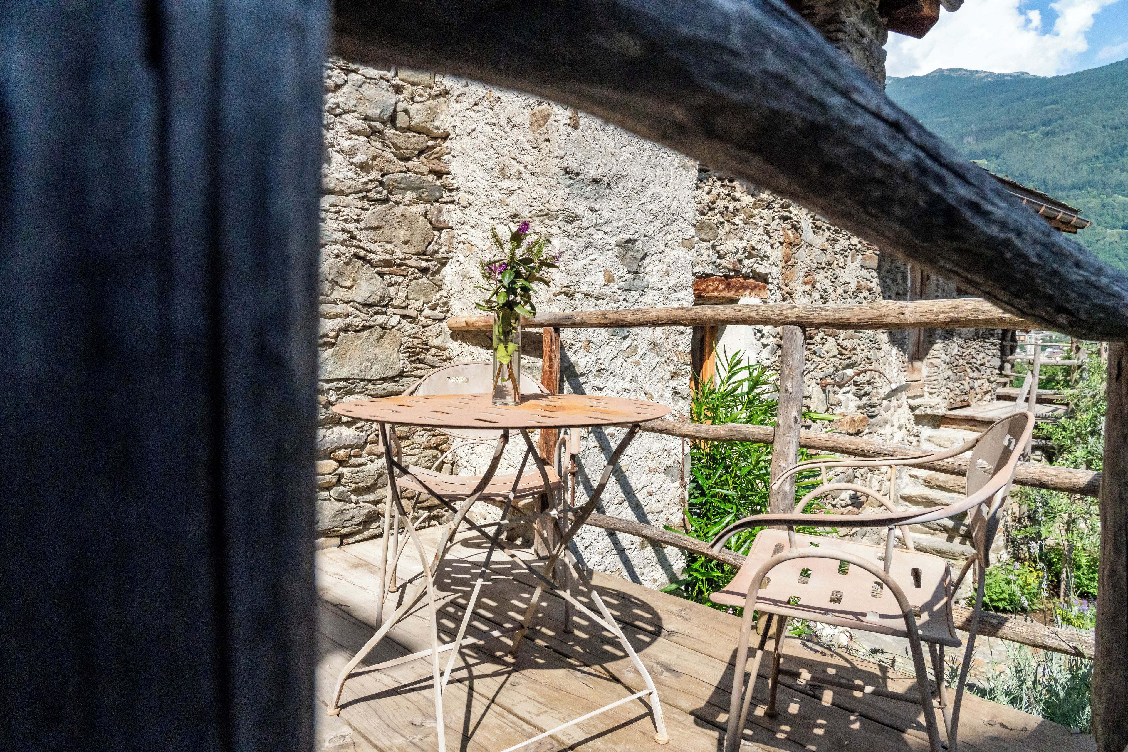 Uma área de estar rústica ao ar livre, com mesa e cadeiras de madeira, cercada por paredes de pedra e vegetação.