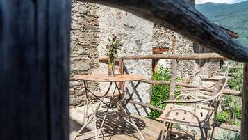 Uma área de estar rústica ao ar livre, com mesa e cadeiras de madeira, cercada por paredes de pedra e vegetação.