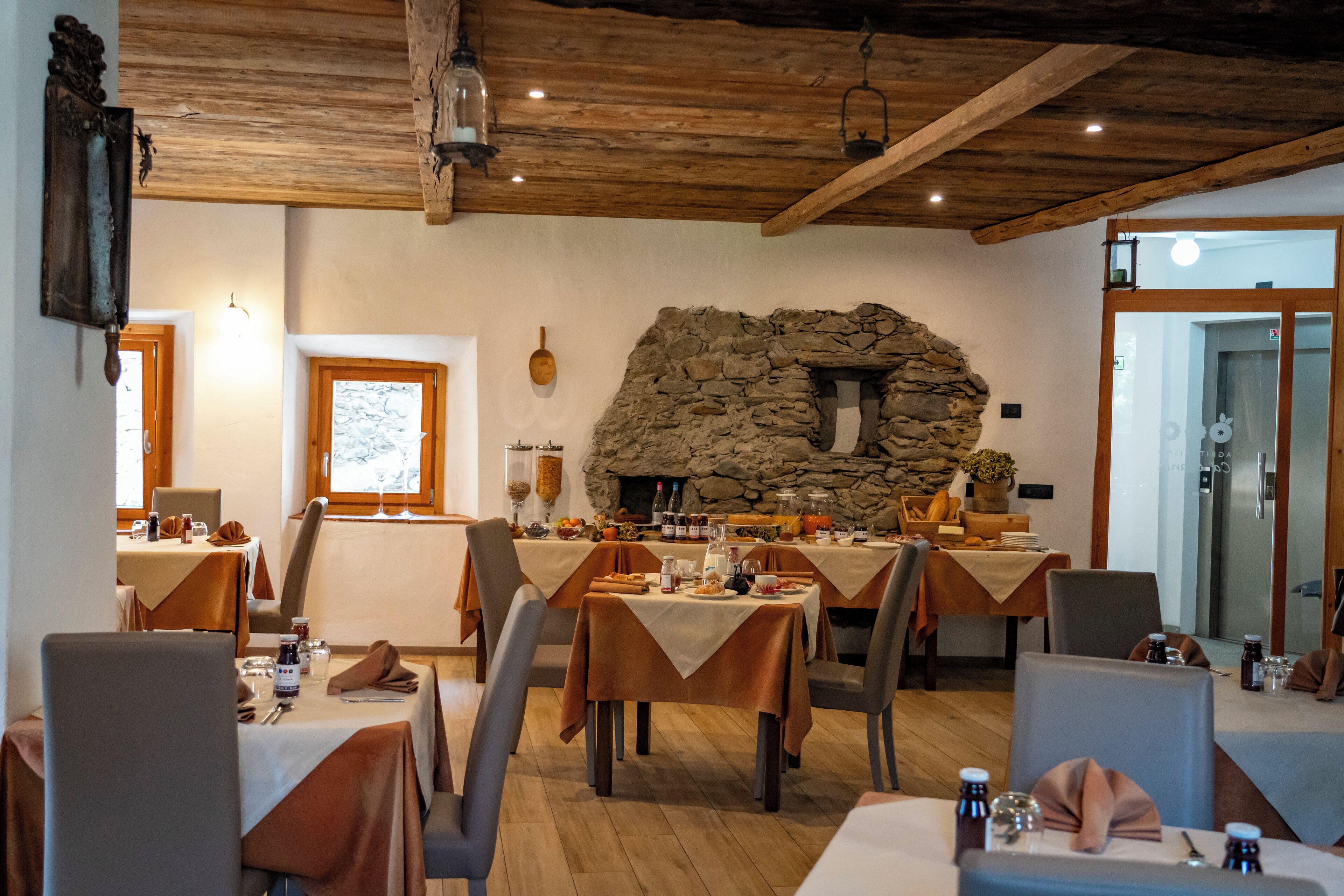 Un comedor con mesas dispuestas para una comida, chimenea de piedra, vigas de madera en el techo y una puerta que da a otra habitación.