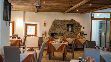 Un comedor con mesas dispuestas para una comida, chimenea de piedra, vigas de madera en el techo y una puerta que da a otra habitación.