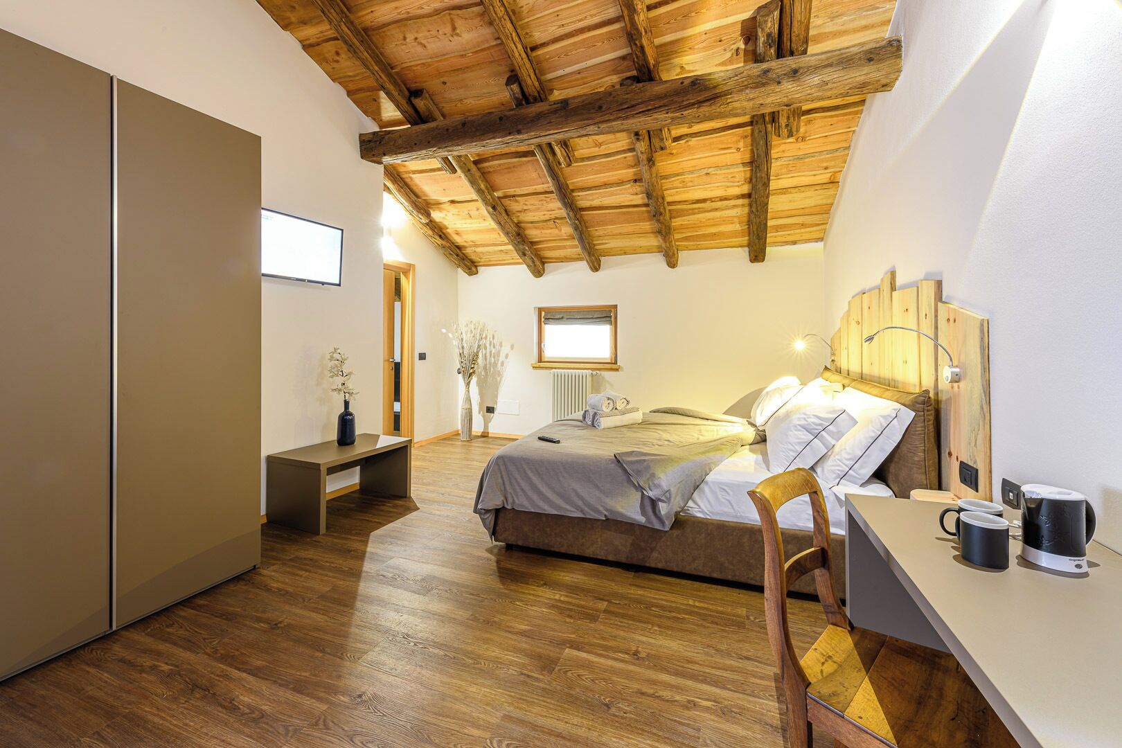 A bedroom with a wooden ceiling, a bed with a gray blanket, a bedside table with a lamp, and a chair.