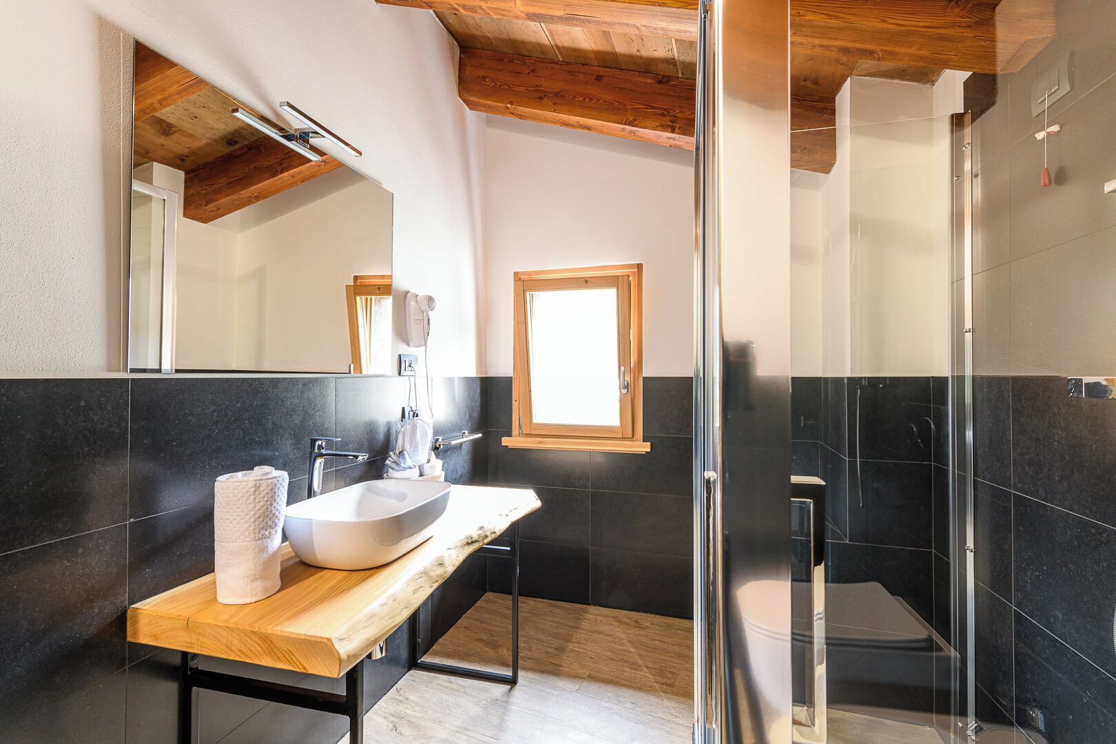 A modern bathroom with a wooden vanity, a large mirror, a glass shower enclosure, and dark tiled walls.