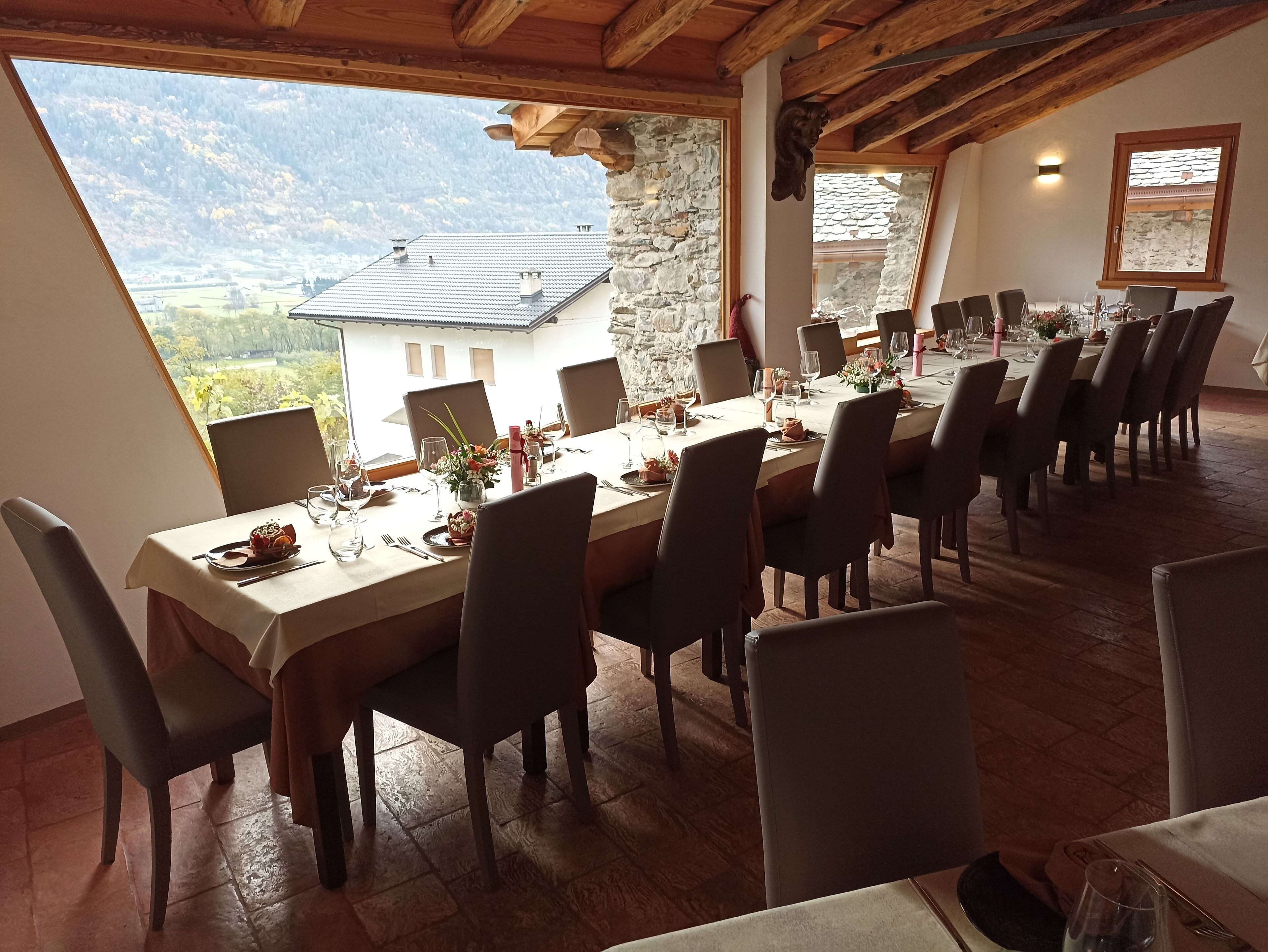 Un'area pranzo con un tavolo lungo apparecchiato per un pasto, travi in legno al soffitto e vista su un paesaggio montano attraverso ampie finestre.