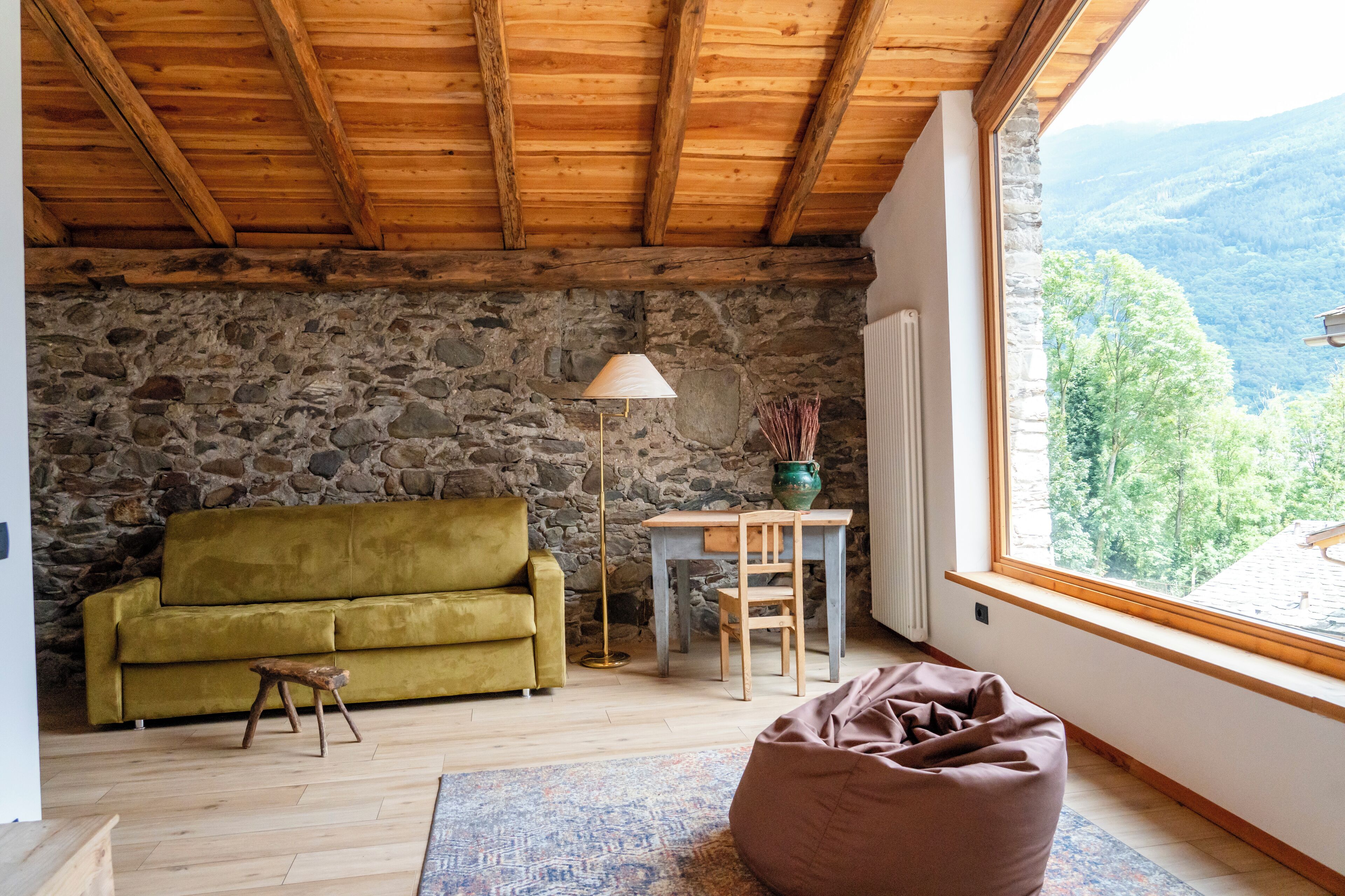 A cozy room with a stone wall, wooden ceiling, a green sofa, a wooden table, a floor lamp, and a view of the mountains.