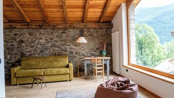 A cozy room with a stone wall, wooden ceiling, a green sofa, a wooden table, a floor lamp, and a view of the mountains.