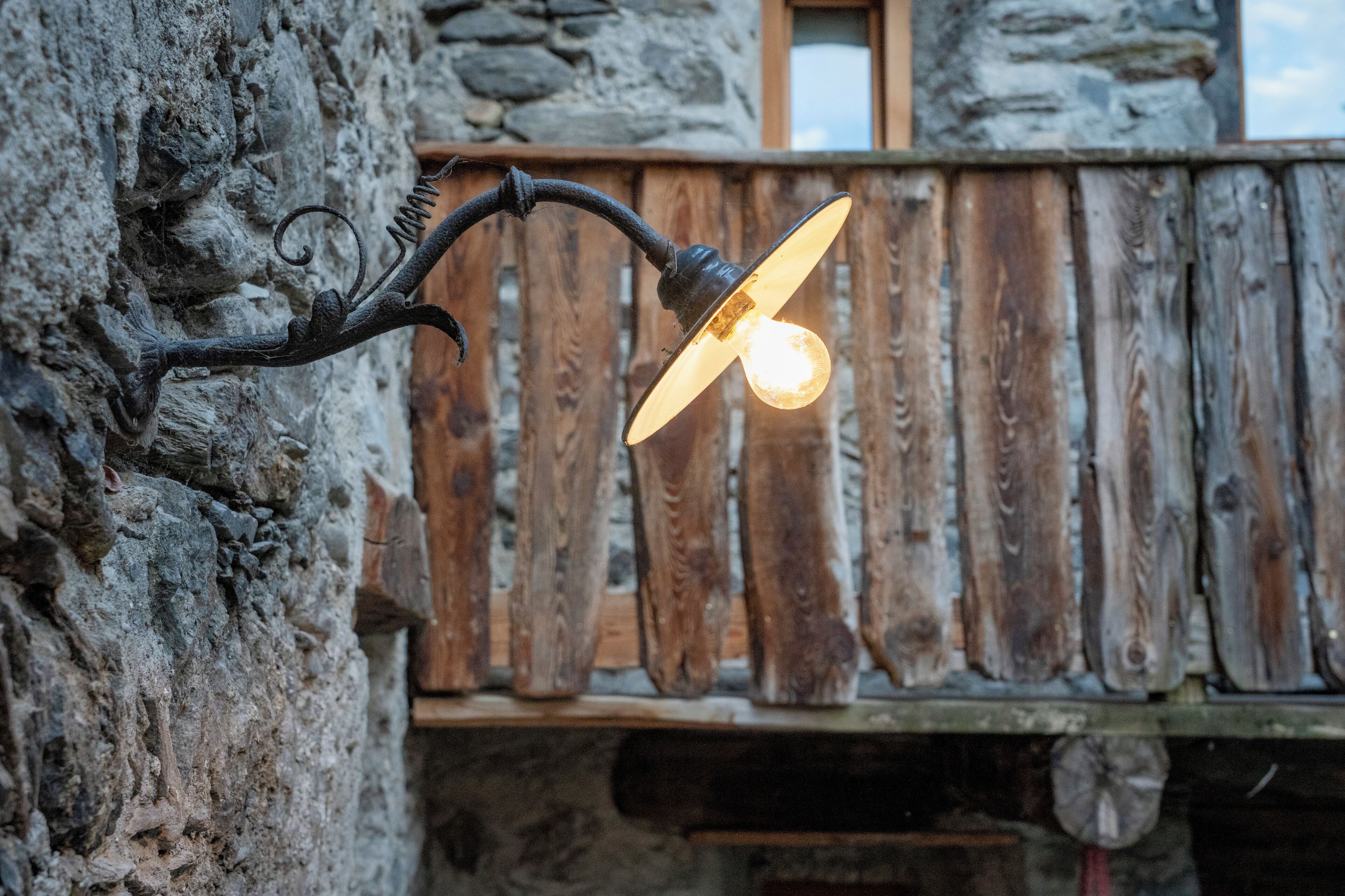 Una lámpara exterior rústica fijada a una pared de piedra, con una valla de madera al fondo.