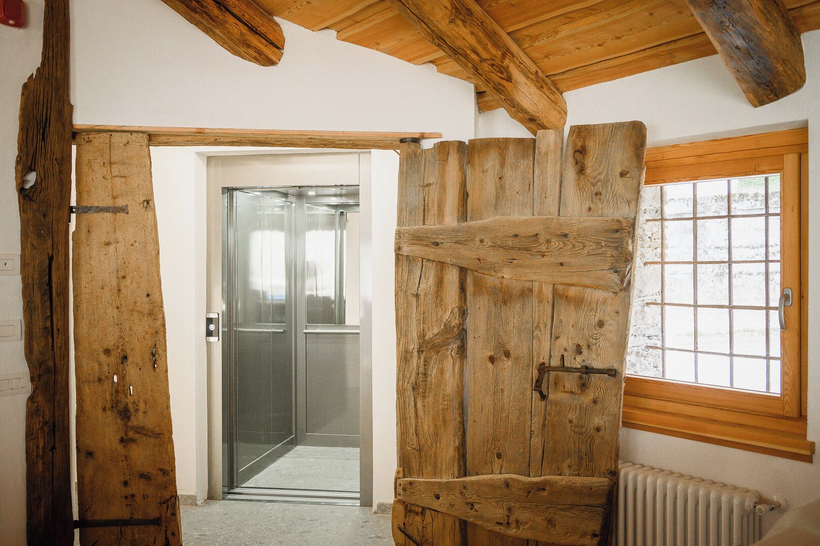 A rustic wooden door leading to a modern elevator, a window with a grid pattern, and a radiator on the wall.