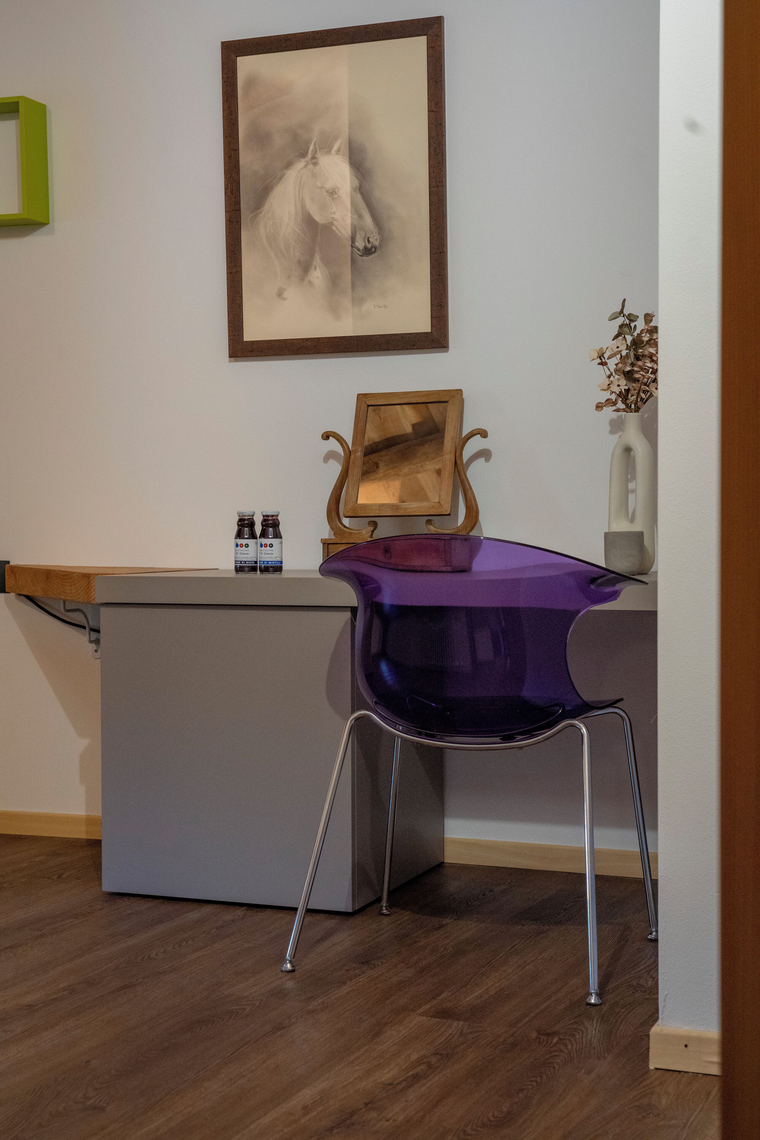 Une pièce comprenant un bureau, une chaise violette, un tableau encadré représentant un cheval, une étagère en bois et un vase contenant des fleurs séchées.
