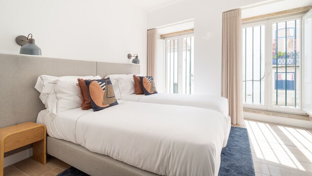 Habitación de hotel con dos camas, una mesita de noche, una lámpara y vistas al exterior a través de amplios ventanales.