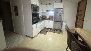 Cocina compacta con gabinetes blancos, microondas, horno y refrigerador. Cuenta con una pequeña mesa de comedor y sillas.