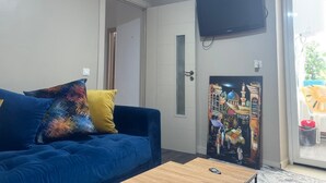 Un salón con un sofá azul, cojines de colores, una mesa de centro de madera, un televisor de pantalla plana montado en la pared y un cuadro colorido sobre una puerta.