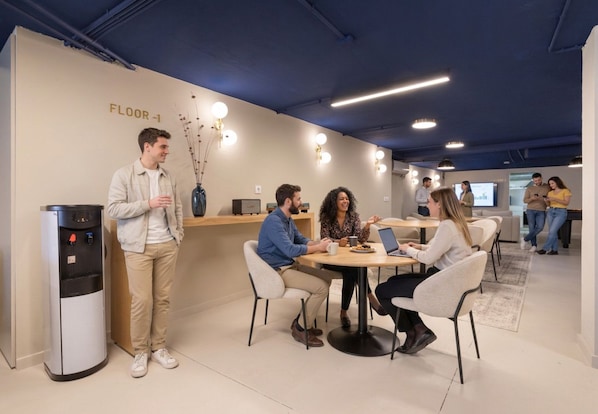Un espacio interior moderno con un dispensador de agua, una mesa redonda con cuatro personas y una pantalla de proyección al fondo.