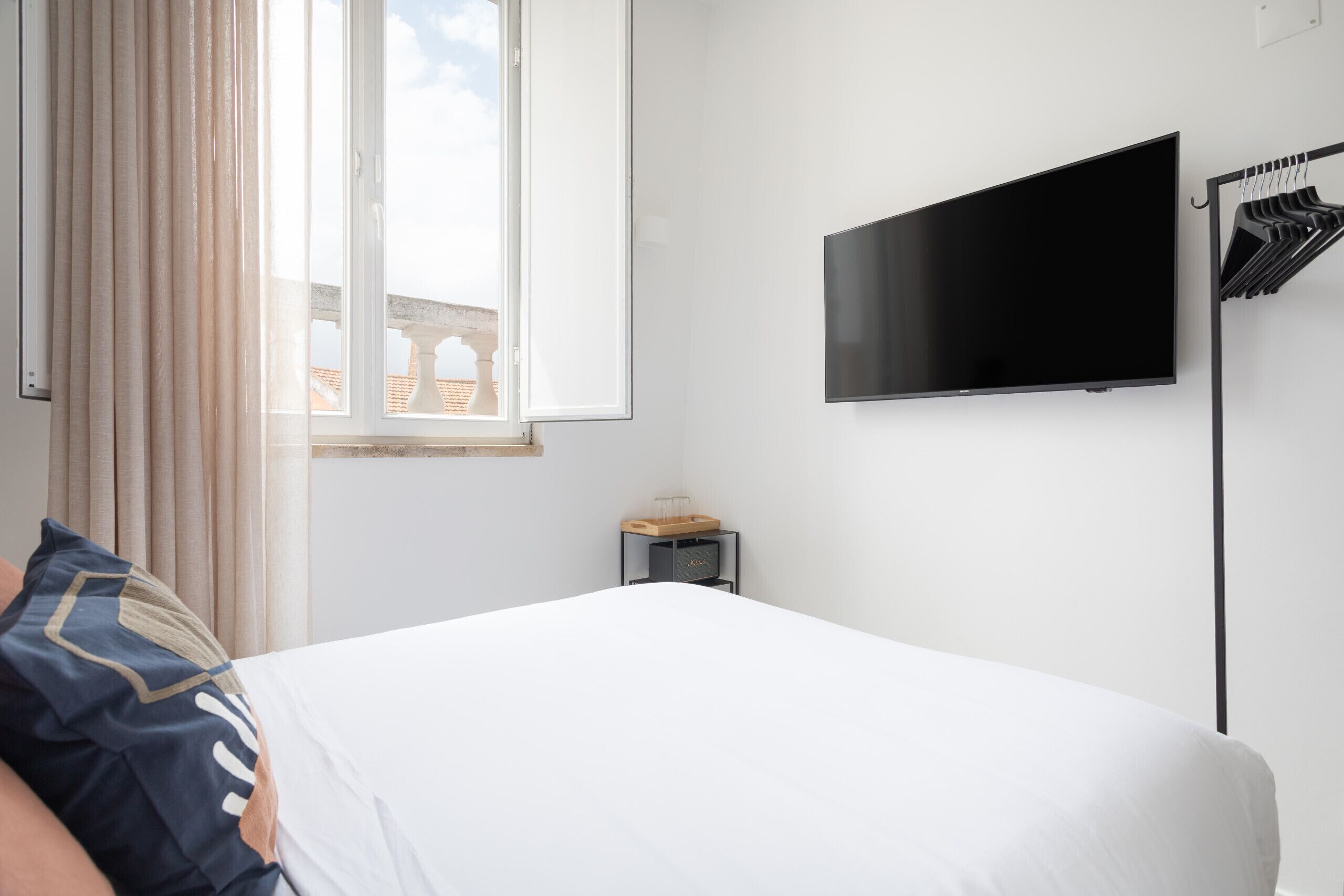 Ein Hotelzimmer mit Bett, Wandfernseher, Fenster mit Vorhängen und einem kleinen Tisch mit Lampe.