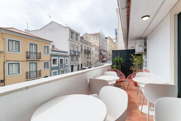 Un balcon avec des tables et des chaises blanches, donnant sur une rue bordée d’immeubles aux couleurs vives.