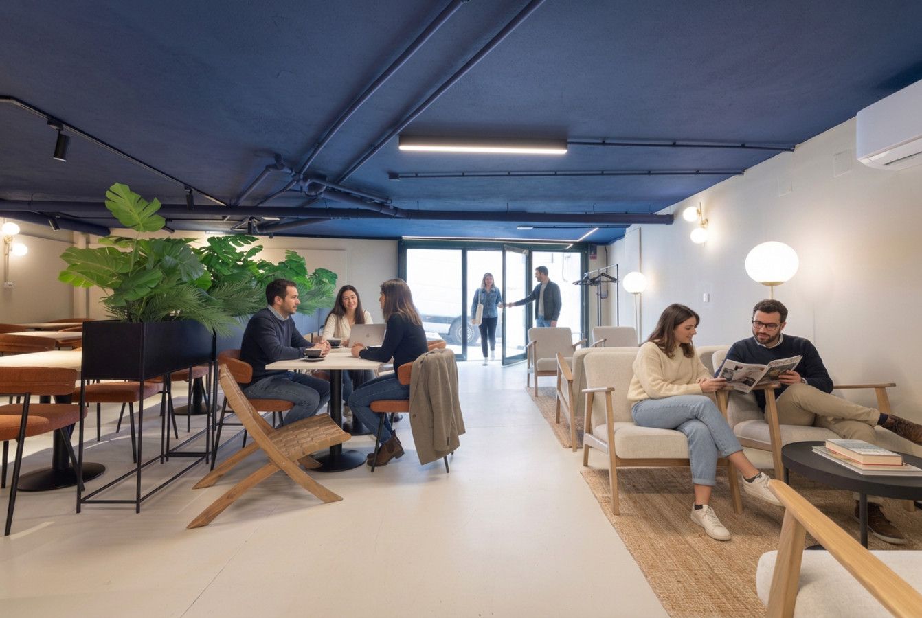 Un espacio de oficina moderno con una variedad de arreglos de asientos, incluyendo mesas y sillas, y una planta grande al fondo.