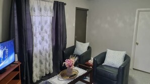 Una sala de estar con dos sofás de cuero negro, una mesa de centro con un florero, un televisor sobre un mueble de madera y una puerta al fondo.