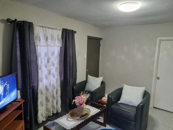 A living room with two black leather sofas, a coffee table with a vase of flowers, a television on a wooden stand, and a door in the background. - Liu guesthouse (Nuku'alofa)